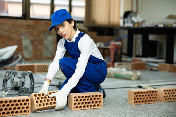 Positive young workwoman in blue overall and cap preparing red bricks for masonry, stacking on construction site indoors..