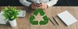© Wanida - Hands of a Business Professional Using Phone with Green Recycle Symbol and Plants on Wooden Desk Background