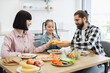 © sofiko14 - Happy Caucasian family with young parents and daughter clinking glasses of juice during breakfast in bright kitchen. Fresh fruits and healthy food create joyful atmosphere.
