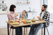 © sofiko14 - Caucasian family with young girl having breakfast with pizza. Parents and child sitting at table enjoying meal. Bright kitchen setting with fruits and vegetables.