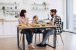 © sofiko14 - Caucasian family with young girl having breakfast with pizza. Parents and child sitting at table enjoying meal. Bright kitchen setting with fruits and vegetables.