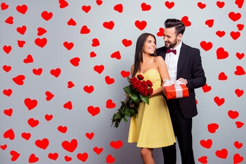 Aufkleber - Portrait with copy space of attractive couple in formal wear, woman in dress holding bouquet of red roses, man in tux embracing her from back side and presenting a packaging red case with gift