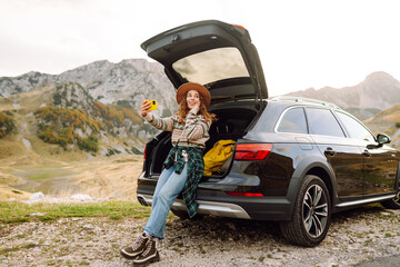 Wall Mural - A young caucasian woman using a smartphone while sitting in the trunk of a car in nature. Travel, weekend, nature, relax and lifestyle concept.