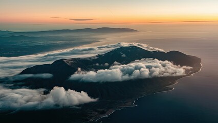 Sticker - Aerial view of a mountain range under soft clouds at sunset with orange hues blending into dark blue sea and distant coastal landscape backdrop