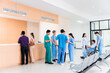 © Rakchanok - Medical staff and patients in a busy hospital reception services at information desk. many patients wait for health care specialists in a hospital waiting area. medical team doctors walking pass lobby