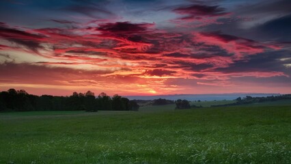 Wall Mural - Vibrant spring sunset over green fields with dramatic red and purple clouds in the sky casting a serene atmosphere at dusk.