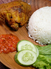 Wall Mural - Closeup traditional Indonesian food consists of white rice, fried chicken, spicy red Sambal, fresh cucumber slices, and lettuce leaves on a wooden plate.