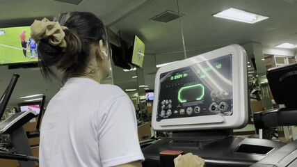 A woman uses a treadmill at a gym. The treadmill has a digital monitor displaying fitness information. She has an earphone in her left ear and her hair is tied back with a scrunchie.