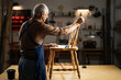 © Andrii Lysenko - Elderly male carpenter working with old chair for restoration in dark carpentry workshop. Furniture restoration, DIY and hobby
