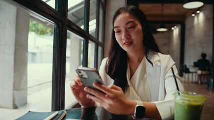 Poster - Professional asian businesswoman enjoying break time, sitting at cafe table, drinking matcha latte and browsing smartphone with relaxed, cheerful demeanor