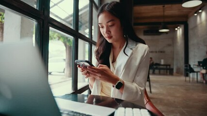 Poster - Young asian businesswoman working remotely, sitting near window in cafe, focusing on smartphone while laptop and keyboard positioned on table, representing modern professional lifestyle