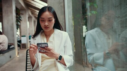 Poster - Professional woman browsing social media on her phone while sitting in a chair inside a modern office building, her reflection visible in the glass wall