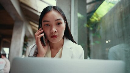 Poster - Professional asian businesswoman sitting in modern office, working on laptop while talking on phone and gesturing with confident expression during business communication