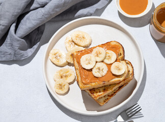 Wall Mural - Peanut butter toast with banana on a plate on a light background with napkin and shadow.