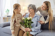 © Studio Romantic - Old grandmother receives congratulations on birthday from daughter and granddaughter poses on sofa with present box and flowers celebrates anniversary. Family time and holiday concept