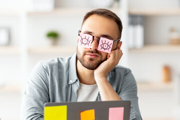 Poster - Workaholism, exhaustion at workplace concept. Tired young man having stickers with open eyes drawing on glasses, sleeping near laptop, pretending to work at home office