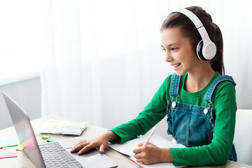 Poster - Studying At Home. Cheerful schoolgirl doing homework and typing on keyboard, sitting at table using laptop, looking at screen, writing essay or making test, taking notes in textbook, free copy space