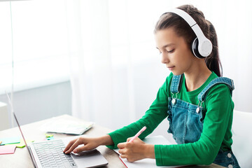 Poster - Educational Lecture Concept. Portrait of diligent teen girl in wireless headphones sitting at table using laptop, writing in copybook taking notes in notebook, listening to teacher, free copy space