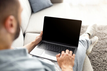 Poster - Unrecognizable young man using laptop on comfy sofa at home, mockup for website design on screen. Millennial guy holding portable pc with copy space, working or studying online