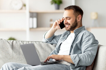 Poster - Attractive young guy talking on smartphone and using laptop computer on couch at home office. Handsome male businessman having phone conversation with client, working on portable pc