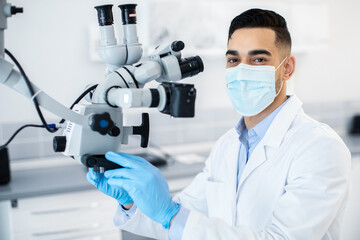 Poster - Arab man dentist wearing protective medical mask using dental microscope in clinic, middle eastern stomatologist doctor working with modern equipment for treating patients, closeup shot