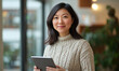 © Keopaserth - woman with long hair is holding tablet while smiling in cozy indoor setting. She wears knitted sweater and stands near window with greenery in background, exuding warm and inviting atmosphere
