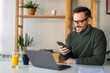 © NDABCREATIVITY - Happy smiling businessman wearing casual clothes and using modern smartphone in his home office
