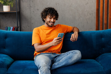 Wall Mural - Young Indian man wear orange casual clothes sits on blue sofa couch hold in hand use mobile cell phone stay at home hotel flat rest relax spend free spare time in living room indoor. Lounge concept.