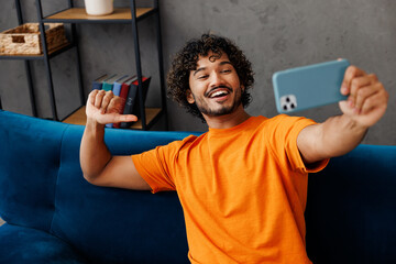 Wall Mural - Young Indian man wear orange casual clothes sits on blue sofa couch doing selfie shot on mobile cell phone stay home hotel flat rest relax spend free spare time in living room indoor. Lounge concept