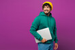 © ViDi Studio - Young happy Indian IT man he wear green hoody yellow hat casual clothes hold closed laptop pc computer look aside on area mock up isolated on plain purple background studio portrait Lifestyle concept