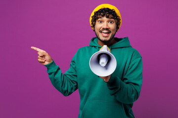 Wall Mural - Young Indian man wear green hoody yellow hat casual clothes hold in hand megaphone scream announces discounts sale Hurry up point finger aside isolated on plain purple background. Lifestyle concept