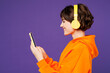 © ViDi Studio - Side view young smiling happy woman she wearing orange hoody casual clothes listen to music in headphones use mobile cell phone isolated on plain purple background studio portrait. Lifestyle concept.