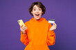 © ViDi Studio - Young woman she wear orange hoody casual clothes using mobile cell phone hold credit bank card doing online shopping order delivery booking tour isolated on plain purple background. Lifestyle concept.