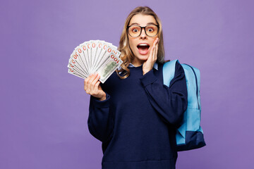 Poster - Young shocked girl student wear blue casual clothes glasses backpack bag hold fan of cash money in dollar banknotes isolated on plain pastel purple background. High school university college concept.