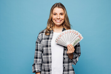 Poster - Young smiling woman she wears checkered grey shirt casual clothes hold in hand fan of cash money in dollar banknotes isolated on plain pastel light blue background studio portrait. Lifestyle concept.