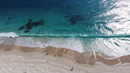 Aufkleber - Sunlight illuminates clear turquoise waves as they gently roll onto a sandy beach, creating a serene and picturesque coastal scene with tourists enjoying the sunny day