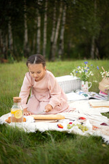 Wall Mural - A gentle summer picnic by the water. Portrait of a blue-eyed girl with long hair in a light dress on the grass and on a beige blanket with flower and fruit. A gentle rest. Warm evening