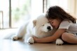 © Milos - In a bright room filled with natural light, a little girl shares a loving embrace with her serene Samoyed, emphasizing the special bond between children and their furry friends.