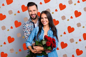 Poster - Portrait of cheerful lovely cute couple with beaming smiles hugging and looking at camera, woman hold a bouquet of red roses on 14 february, celebrating anniversary over grey background