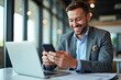 © SimpleDesignStudio - A Smiling Businessman Engaged in Work, Texting on His Phone While Using a Laptop in a Modern Office Environment Filled with Natural Light and Contemporary Decor