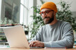 © Maria Vitkovska - Smiling bearded man wearing glasses and beanie using laptop in bright office