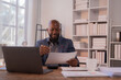 © Tj - Businessman is smiling while reviewing documents and working on a laptop in a modern office, suggesting productivity and focus in a professional environment