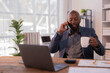 © Tj - Businessman sitting at a modern desk, engaging in a phone conversation while sipping coffee. A laptop, calculator, and documents are spread out, highlighting a busy morning routine