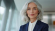© HavingFunShop - A senior businesswoman with shoulder-length silver hair, dressed in a tailored navy-blue jacket and a white blouse, standing in front of a softly lit modern office space