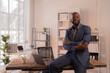 © Tj - Smiling black entrepreneur sitting on his desk with crossed arms in a luminous office, representing business success, leadership and confidence in a corporate environment