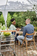 © Seventyfour - Vertical shot of female architect of Black ethnicity talking to male professional landscaper sourcing plants for projects discussing wholesale deal, while sitting at table with laptop in garden