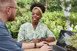 © Seventyfour - Medium shot of smiling African American female landscape designer with phone in hands discussing garden project with male colleague, while sitting at table with laptop in sunlit garden