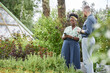 © Seventyfour - Positive African American female landscape designer using mobile phone while discussing with male client garden project providing man with professional assistance captured against lively greenery