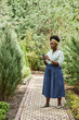 © Seventyfour - Vertical full length of African American female landscape designer using mobile phone managing business while walking along lively green alley of plant nursery, copy space