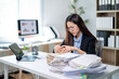 © Wasana - A woman is sitting at a desk with a pile of papers in front of her
